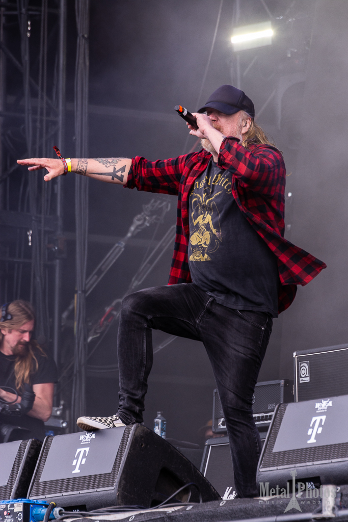 A male singer with long hair wearing a black cap, a black graphic t-shirt, and a red plaid shirt performs energetically on stage, microphone in hand. In the background, a musician is operating a camera, with amplifiers visible.